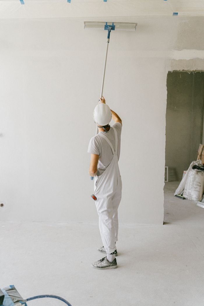Back view of a painter in overalls and hard hat painting an interior wall with a roller.